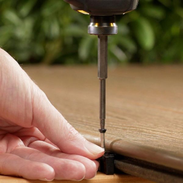 A person's hand is shown holding a small black bracket while a drill drives a screw into a wooden plank. The setting appears to be a woodworking or DIY project, with a natural wood texture and green foliage blurred in the background. The image focuses on the precision and care involved in the assembly process. No visible text or numbers are present.