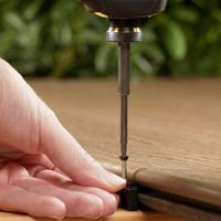 A person's hand is shown holding a small black bracket while a drill drives a screw into a wooden plank. The setting appears to be a woodworking or DIY project, with a natural wood texture and green foliage blurred in the background. The image focuses on the precision and care involved in the assembly process. No visible text or numbers are present.