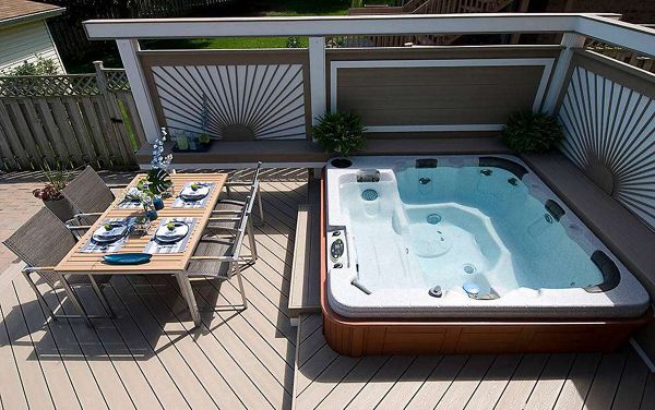 A contemporary outdoor deck features a large built-in hot tub filled with clear water. Adjacent to the hot tub is a dining table set for six, complete with plates, glasses, and napkins, suggesting a meal or gathering. The deck is surrounded by decorative privacy panels and potted plants, creating a relaxed and inviting atmosphere. The setting is bright and clean, with neutral tones and geometric patterns.