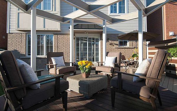 A contemporary backyard patio features cushioned outdoor chairs arranged around a central table with a yellow flower arrangement. The setting is shaded by a geometric pergola structure, with a house and large windows in the background. The scene is well-lit, evoking a relaxed and inviting atmosphere, with neutral and earthy tones dominating the palette. No visible people or text are present in the image.