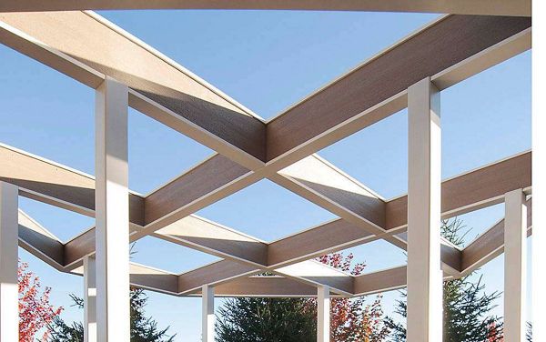 The image features a contemporary pergola with intersecting beams forming a geometric pattern overhead. The setting is outdoors, with clear blue sky visible through the open roof and trees in the background. The structure's clean lines and neutral palette create a modern, architectural mood. No visible text, logos, or numeric values are present in the image.
