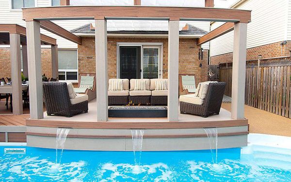 A contemporary outdoor patio features cushioned wicker seating arranged around a rectangular fire pit. The setting overlooks a bright blue swimming pool with three small waterfalls cascading from the deck. The space is framed by a wooden pergola and bordered by a suburban house and privacy fence, creating a relaxing and inviting atmosphere.