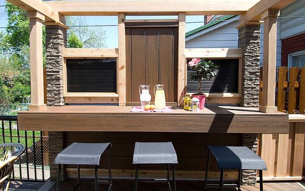 A modern outdoor patio bar features three black stools lined up at a wooden counter. On the bar, there are two clear pitchers filled with lemonade and water, along with glasses and a small pink napkin holder. The setting is bright and sunny, with stone pillars and wooden beams framing the space, and greenery visible in the background.