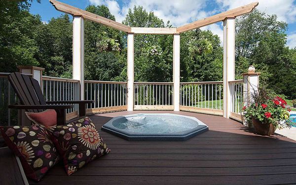 A modern outdoor deck features a built-in hot tub surrounded by wooden railings and lush greenery. Two colorful patterned pillows rest on a wooden chair, adding a cozy touch to the seating area. The setting is bright and inviting, with natural sunlight and a clear blue sky. The overall mood is relaxing and tranquil, ideal for leisure and outdoor enjoyment.
