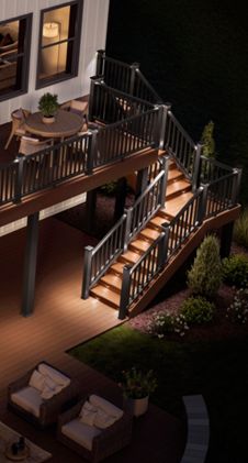 Elevated wooden deck extends from a modern house, illuminated by warm accent lights along the railings and stairs. The scene is set at night, highlighting the outdoor living space with a dining table on the upper deck and a seating area below. Stairs connect the two levels, and soft landscape lighting reveals surrounding plants and garden beds. Large windows and glass doors on the house reflect the cozy ambiance of the outdoor area.