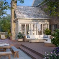 A spacious outdoor patio featuring wicker furniture, a wooden dining table, and a stone fireplace. The setting includes lush greenery, a well-maintained deck, and a charming house exterior with large windows. The scene is ideal for gatherings or relaxation in a serene environment.