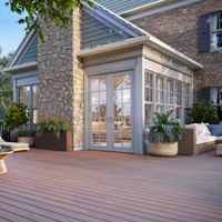 A spacious wooden patio featuring wicker chairs and lush greenery. The setting includes a stone and brick house with large glass doors leading to the interior. Decorative planters and soft lighting create a serene and inviting atmosphere.