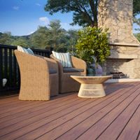 A spacious outdoor patio featuring wicker chairs with striped cushions and a modern fire pit table. The setting includes a stone fireplace surrounded by lush greenery and trees, creating a serene atmosphere. Wooden deck flooring adds warmth to the scene, complemented by lounge chairs and a scenic backdrop.
