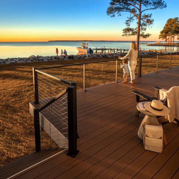 A woman and a young child stand on a wooden deck overlooking a tranquil lake at sunset. The scene features a clear sky, calm water, and a dock with people and a boat in the distance. Warm golden light bathes the landscape, creating a peaceful and inviting atmosphere. Outdoor furniture and a hat are visible on the deck, enhancing the relaxed mood.