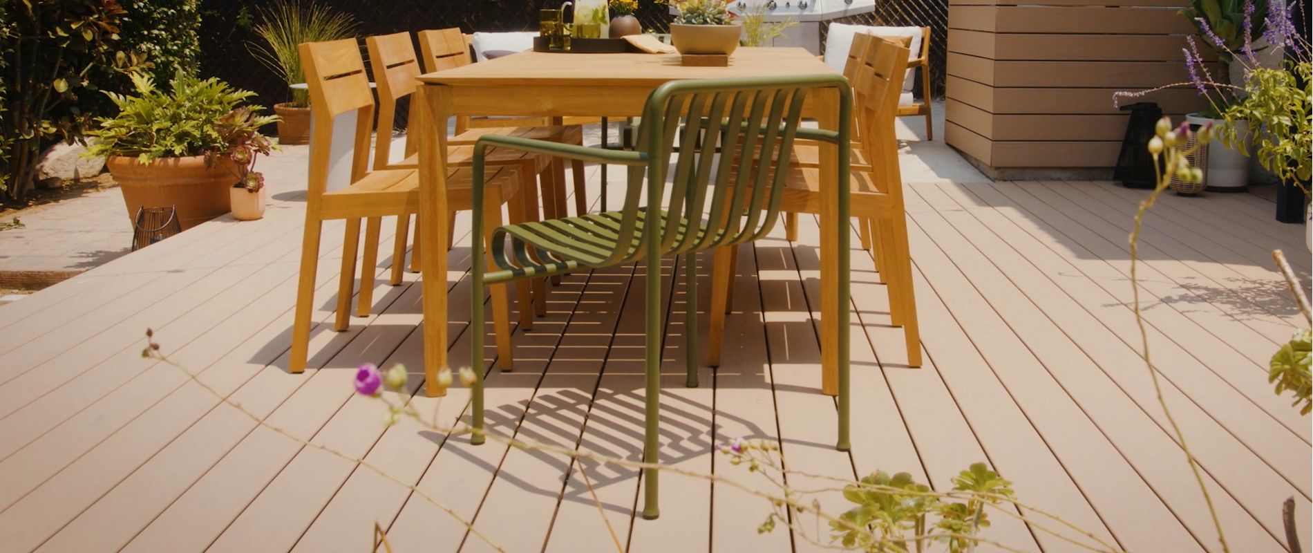 A modern outdoor dining table with several wooden chairs and one green metal chair is arranged on a spacious wooden deck. The setting is a sunlit backyard patio surrounded by plants, potted flowers, and a barbecue grill in the background. The scene conveys a relaxed, inviting atmosphere with natural tones and greenery. No visible text or numbers are present in the image.