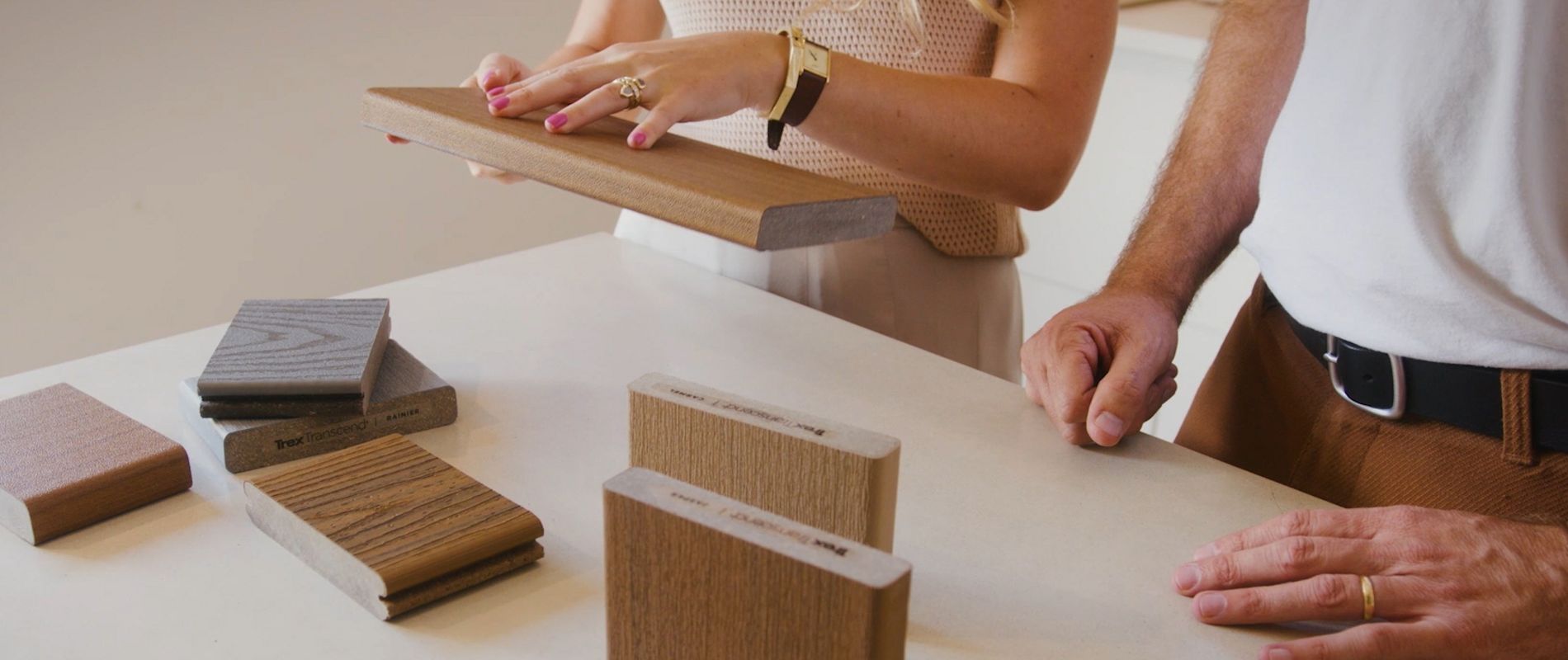 Two adults are closely inspecting various rectangular wood samples laid out on a light-colored table. The setting appears to be indoors, possibly a design studio or showroom, with a focus on material selection. The individuals' hands are visible, highlighting engagement with the textures and finishes of the wood pieces. The overall mood is professional and collaborative, with neutral and warm tones dominating the palette.