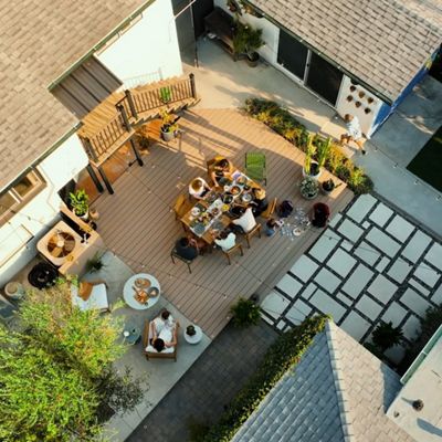 This image captures an overhead perspective of a group of people enjoying a meal together on a spacious wooden deck in a residential backyard. The setting features modern outdoor furniture, a mix of greenery, and paved stone pathways. The gathering appears relaxed and social, with food and drinks arranged on the central table. The color palette is warm and natural, with sunlight casting soft shadows across the scene.