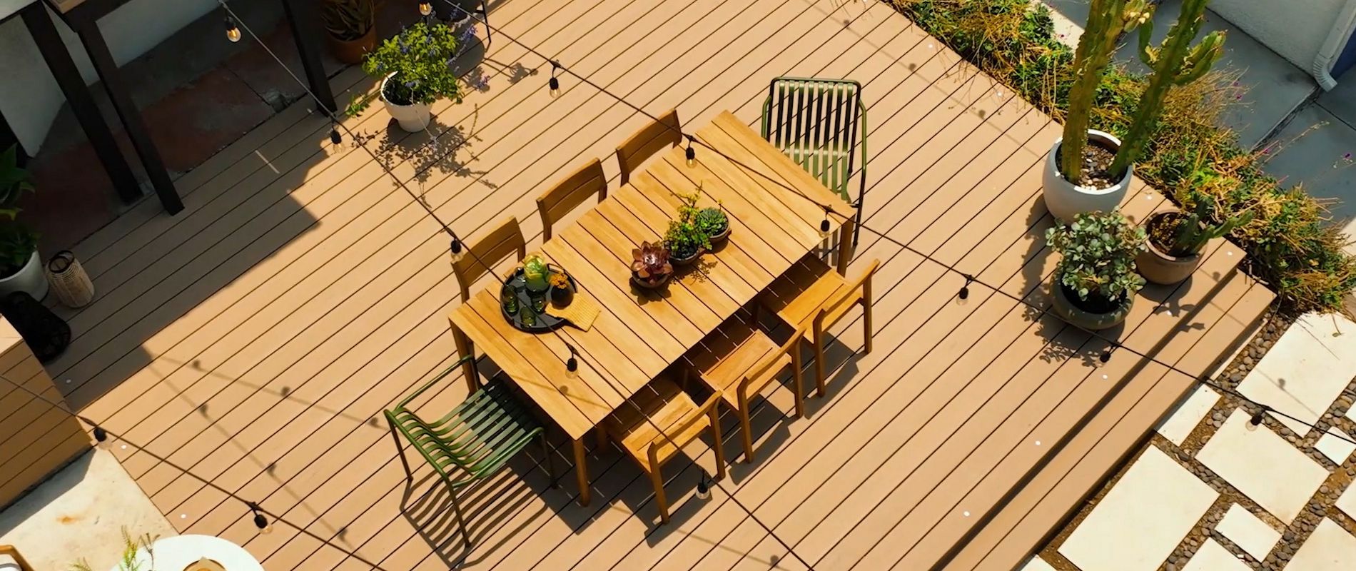 A contemporary outdoor patio features a wooden deck with a rectangular dining table and six chairs. The setting is decorated with potted plants, string lights, and a mix of green and natural tones. The area is surrounded by garden beds and stone pavers, creating a relaxed and inviting atmosphere. The image is taken from an overhead perspective, highlighting the clean lines and organized layout.