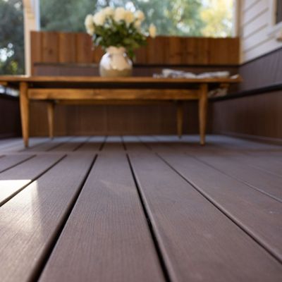 A close-up view of a brown wooden patio deck is shown, leading to a wooden table with a white vase of flowers. The setting appears to be an outdoor porch or terrace, with sunlight filtering through trees in the background. The mood is warm and inviting, with natural tones and a cozy atmosphere. No visible text or numbers are present in the image.