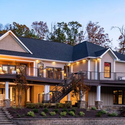 A spacious, contemporary two-story house is shown with a wraparound balcony and exterior lighting illuminating the structure at dusk. The home features large windows, a stone chimney, and an outdoor staircase leading to the upper level. The setting includes landscaped gardens and mature trees with autumn foliage, creating a warm and inviting atmosphere.