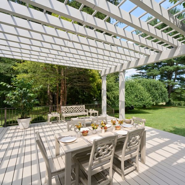 An outdoor dining area sits beneath a large white pergola on a spacious backyard deck. A long table with matching chairs is set with plates and small floral arrangements, ready for a meal. A decorative bench and potted plant add detail to the seating area. Lush green lawn, trees, and shrubs surround the deck, creating a peaceful garden setting under a bright blue sky.