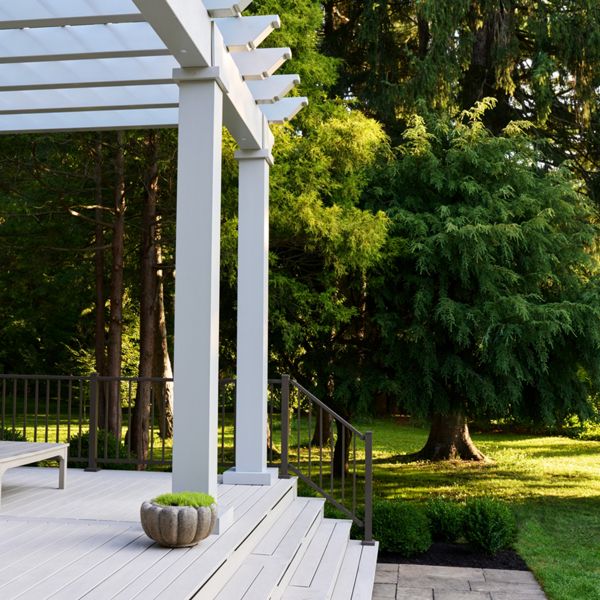 A modern white pergola and deck extends from a house into a lush backyard. The scene shows clean steps leading down to a stone patio bordered by neatly trimmed shrubs. A small decorative planter sits on the deck, adding a touch of greenery. Tall evergreen trees and a sunlit lawn create a peaceful, natural backdrop.
