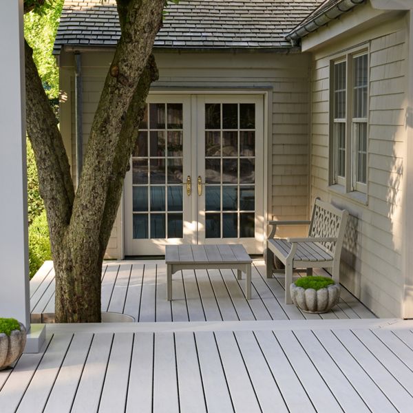 A light-colored wooden deck extends from the back of a house, leading to glass-paneled French doors. A small bench and matching table create a simple seating area beneath the shade of a large tree. Potted plants with green foliage add a touch of color to the neutral exterior siding. The scene suggests a quiet, inviting outdoor space for relaxation in a suburban yard.