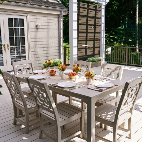 A spacious wooden deck features a large outdoor dining table set for a meal. Neutral-toned chairs surround the table, which is decorated with plates, glasses, and colorful floral centerpieces. The deck extends toward a railing overlooking lush green trees, creating a serene backyard setting. Additional seating and tables are visible in the background, suggesting space for entertaining and gatherings.