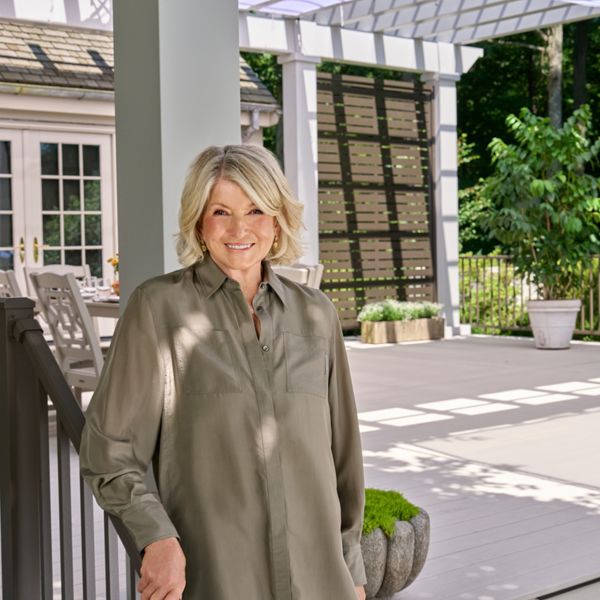 A smiling middle-aged woman stands on the edge of a spacious outdoor deck. She leans casually against a railing in front of a large house with many windows and French doors. The patio features pergola structures, potted plants, and outdoor seating under soft daylight. Lush green trees surround the space, creating a serene backyard atmosphere.