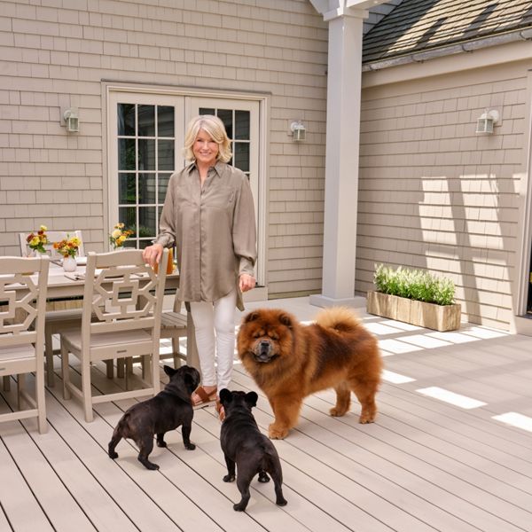 A mature woman stands on a light-colored deck with three dogs, including a fluffy large dog and two small black dogs. She is positioned near an outdoor dining table set with chairs and vases of flowers. The scene is set against a shingled house exterior with large windows and a glass door. Soft natural light and neutral tones create a calm, welcoming atmosphere.