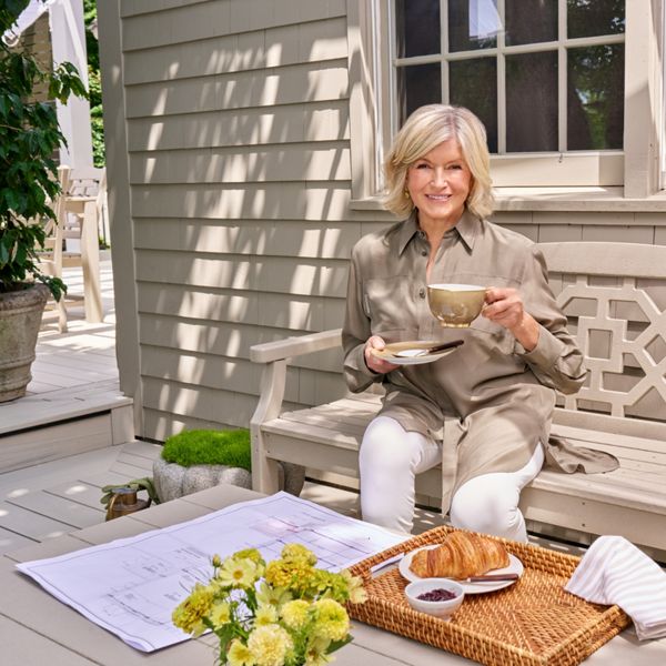 A woman sits on a light-colored wooden bench on a bright outdoor patio, holding a cup and saucer of coffee or tea. She is dressed in a neutral-toned shirt and white pants, smiling toward the camera. In front of her, a wicker tray holds a croissant, small dish of jam, and a striped napkin. Architectural drawings or plans and a small bouquet of yellow flowers rest on the bench beside her, while potted greenery decorates the background.