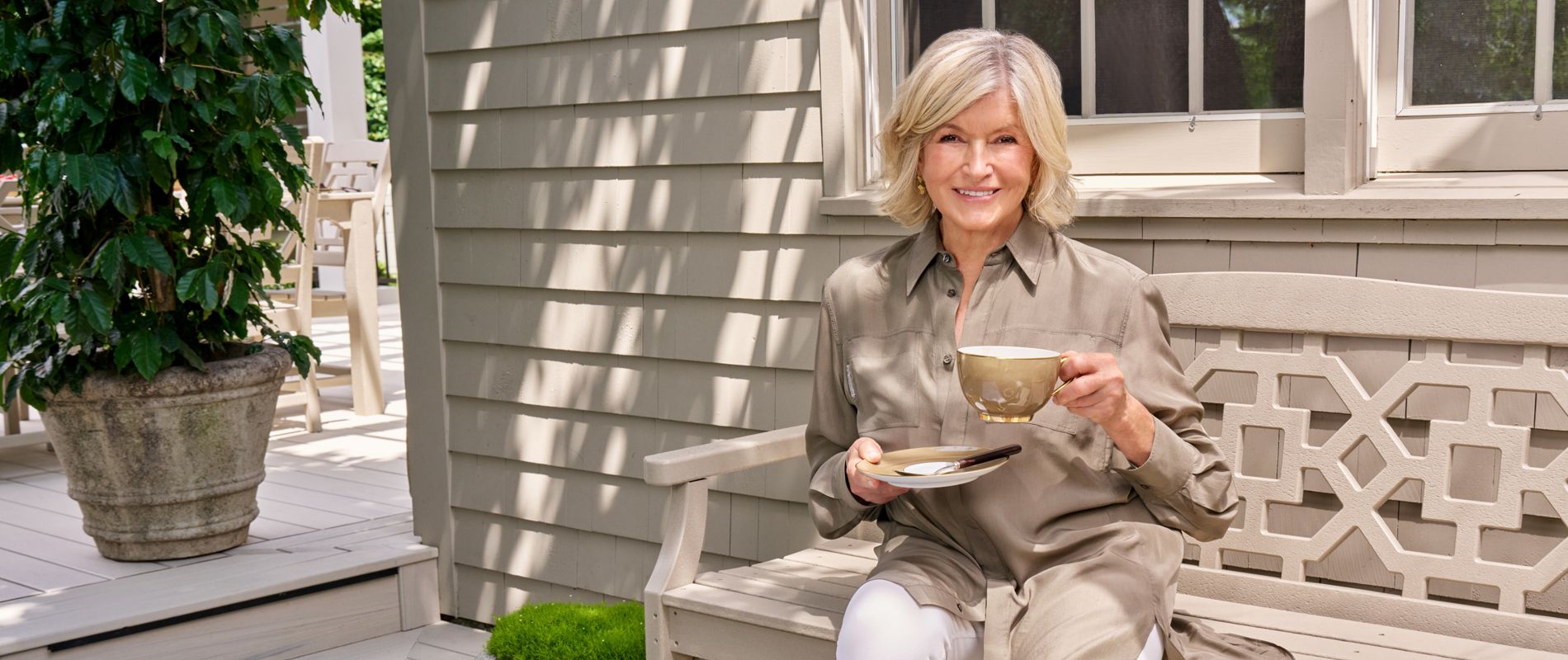 A woman sits on a light-colored wooden bench on a bright outdoor patio, holding a cup and saucer of coffee or tea. She is dressed in a neutral-toned shirt and white pants, smiling toward the camera. In front of her, a wicker tray holds a croissant, small dish of jam, and a striped napkin. Architectural drawings or plans and a small bouquet of yellow flowers rest on the bench beside her, while potted greenery decorates the background.