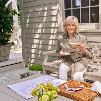 A woman sits on a light-colored wooden bench on a bright outdoor patio, holding a cup and saucer of coffee or tea. She is dressed in a neutral-toned shirt and white pants, smiling toward the camera. In front of her, a wicker tray holds a croissant, small dish of jam, and a striped napkin. Architectural drawings or plans and a small bouquet of yellow flowers rest on the bench beside her, while potted greenery decorates the background.