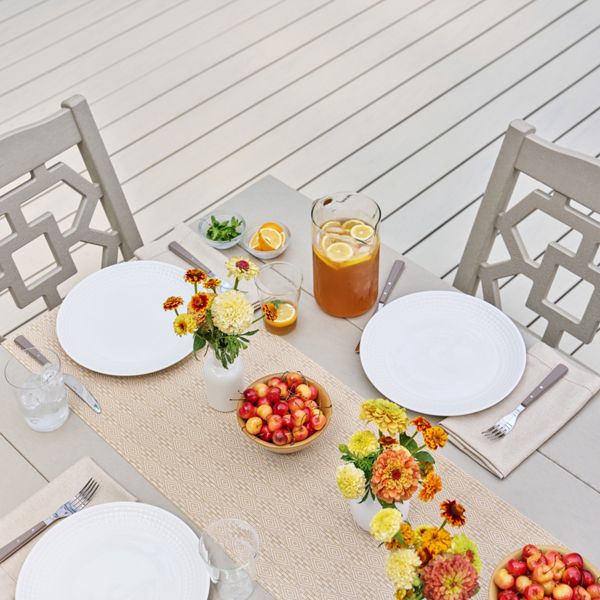 An outdoor dining table is set for a casual summer meal on a light-colored patio deck. Two white plates with cloth napkins and cutlery are arranged on either side of a woven table runner. A pitcher and glasses of iced tea with citrus slices sit alongside small bowls of fresh cherries. Colorful floral arrangements in small vases run down the center of the table, adding bright yellow and orange accents. The scene feels airy, relaxed, and ready for an al fresco gathering.
