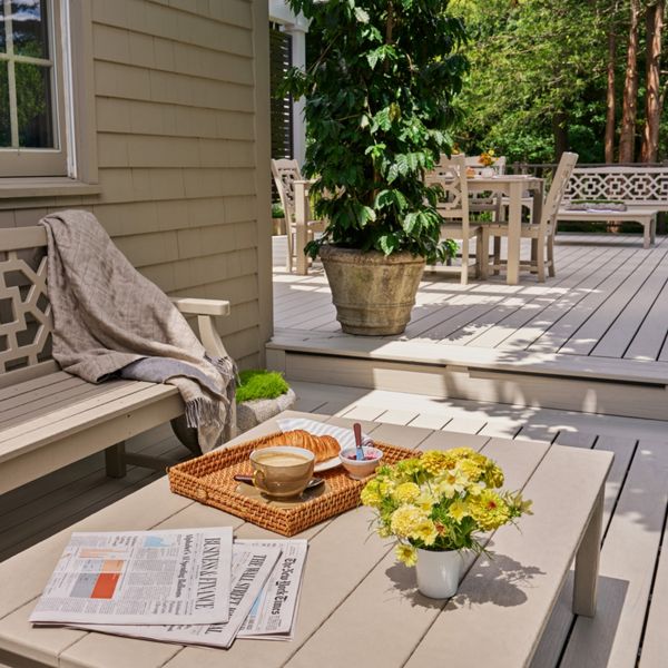 A bright outdoor deck features a bench and low table arranged for a quiet morning. A tray with a coffee cup, bowl, and spoon sits on the table beside a newspaper and a small vase of yellow flowers. In the background, a potted plant and additional patio furniture create a layered, inviting space. Soft neutral tones and natural greenery contribute to a calm, relaxed atmosphere.