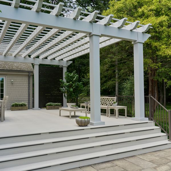 A spacious white pergola covers an elevated backyard deck attached to a house. The deck features outdoor dining furniture, a decorative bench, and potted plants arranged neatly along the edges. Wide steps lead down to a stone patio and a lush green lawn surrounded by trees. Soft neutral tones of the structure contrast with the vibrant greenery, creating a calm and inviting outdoor living space.
