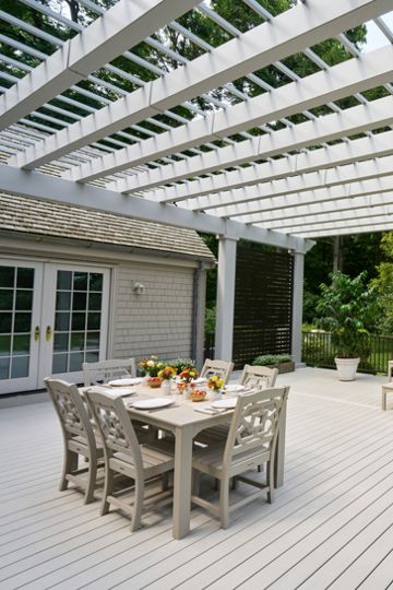 A large white wooden deck extends from a house into a lush backyard. A white pergola structure covers part of the deck, creating an inviting outdoor living space. A dining table is set with dishes and drinks, while a bench and coffee table form a separate seating area. Surrounding trees and greenery provide a serene, private garden atmosphere.