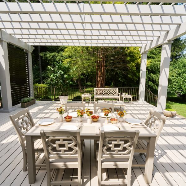 An elegant outdoor dining table is set beneath a modern white pergola on a spacious deck. The table is arranged with plates, glasses, and small floral centerpieces, ready for a meal. Decorative chairs and a matching bench add a cohesive design to the seating area. Lush green trees and shrubs surround the deck, creating a serene garden backdrop with dappled sunlight and shadows from the pergola overhead.