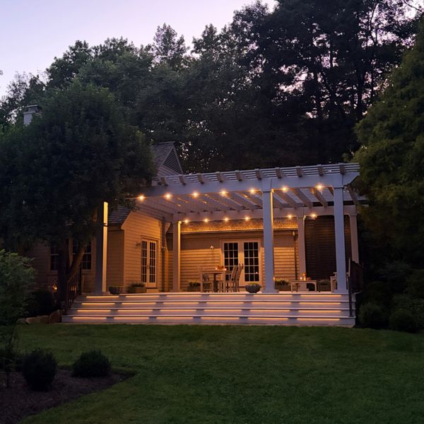 A spacious backyard patio features a white pergola attached to a house at dusk. Warm string lights illuminate the pergola and the wide steps leading down to a manicured lawn. Outdoor furniture is arranged beneath the structure, creating a cozy seating area. Tall trees and shrubs surround the yard, adding privacy and a tranquil atmosphere under the twilight sky.
