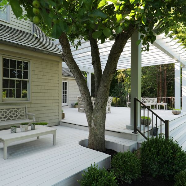 A spacious white wooden deck extends from a traditional house into a landscaped backyard. A large tree grows through the deck, providing natural shade near a seating area with benches and a small table. A raised section with steps leads to a pergola structure supported by columns. Neatly trimmed shrubs and stone pavers frame the outdoor living space, creating a calm and inviting atmosphere.