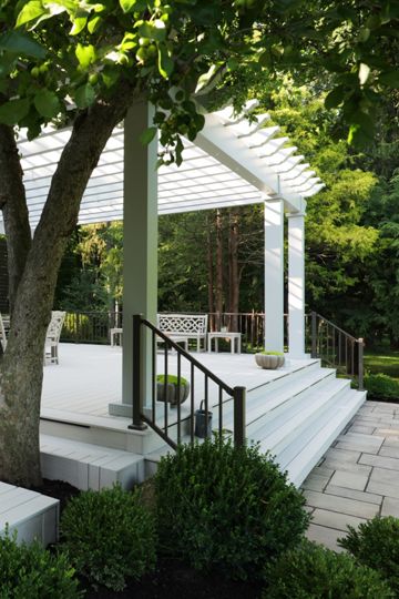 A spacious white wooden deck extends from a traditional house into a landscaped backyard. A large tree grows through the deck, providing natural shade near a seating area with benches and a small table. A raised section with steps leads to a pergola structure supported by columns. Neatly trimmed shrubs and stone pavers frame the outdoor living space, creating a calm and inviting atmosphere.