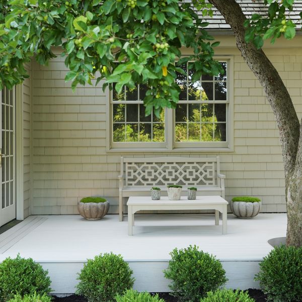 A serene backyard patio features a decorative wooden bench and matching coffee table placed against a light-colored shingled house. Potted plants sit symmetrically on either side of the bench, adding greenery to the clean white deck. A large tree in the foreground provides shade, while manicured shrubs line the edge of the patio. French doors and multi-pane windows enhance the cozy, inviting outdoor living space.