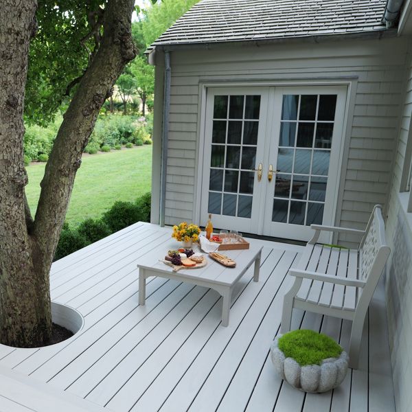 A bright white wooden deck wraps around a large tree beside a house with French doors. A simple outdoor table is set with food and drinks, creating a casual dining area. A bench and a small green cushion add seating and a pop of color. Lush green lawn and shrubs extend into the background, giving the space a peaceful garden atmosphere.