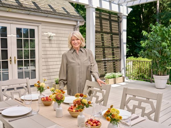 A woman stands beside a neatly arranged outdoor dining table on a patio. The table is set with white plates, glassware, and small vases filled with yellow and orange flowers. Bowls of fresh fruit add color to the neutral-toned furniture and decor. A light-colored house and lush green trees frame the relaxed garden setting.