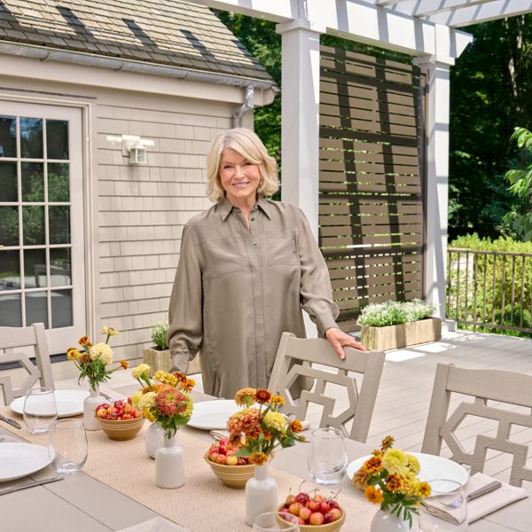 A woman stands beside a neatly arranged outdoor dining table on a patio. The table is set with white plates, glassware, and small vases filled with yellow and orange flowers. Bowls of fresh fruit add color to the neutral-toned furniture and decor. A light-colored house and lush green trees frame the relaxed garden setting.