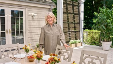A woman stands beside a neatly arranged outdoor dining table on a patio. The table is set with white plates, glassware, and small vases filled with yellow and orange flowers. Bowls of fresh fruit add color to the neutral-toned furniture and decor. A light-colored house and lush green trees frame the relaxed garden setting.
