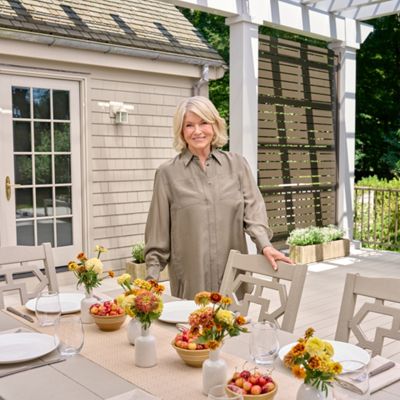 A woman stands beside a neatly arranged outdoor dining table on a patio. The table is set with white plates, glassware, and small vases filled with yellow and orange flowers. Bowls of fresh fruit add color to the neutral-toned furniture and decor. A light-colored house and lush green trees frame the relaxed garden setting.