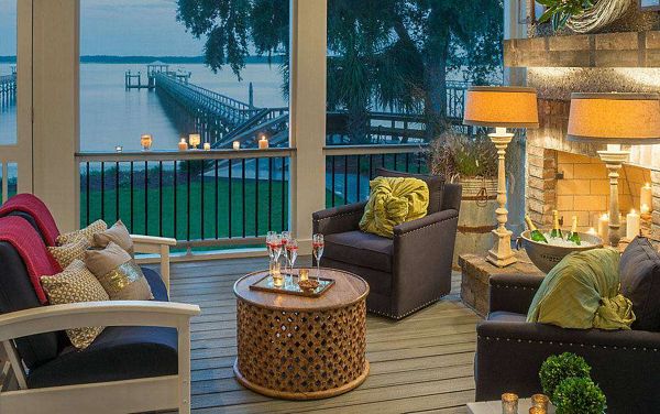 A stylish outdoor patio overlooks a serene waterfront with a long dock stretching into the distance. The seating area features cushioned chairs, a round wooden table, and decorative pillows, all illuminated by warm lamp light and numerous candles. The tranquil setting is enhanced by the soft glow of dusk, creating a relaxing and inviting atmosphere.
