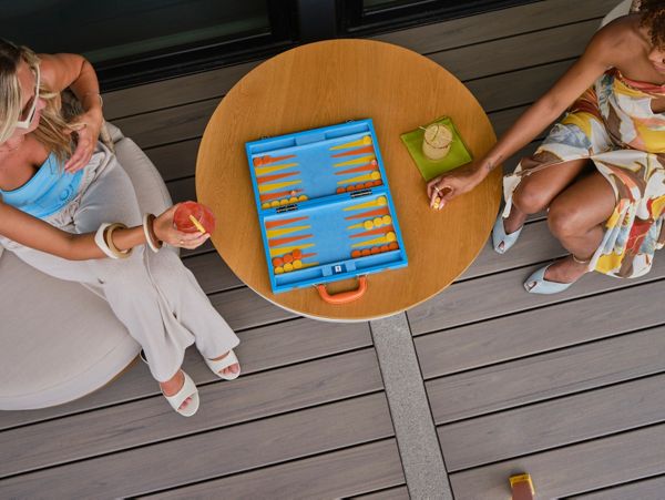 Two women are seated at a round wooden table, engaged in a game of backgammon. The setting is an outdoor patio with modern decking, and each woman has a drink nearby. The scene is bright and relaxed, with summery attire and a casual atmosphere. The backgammon set is blue with orange and yellow pieces, adding a pop of color to the composition.