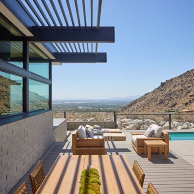 A contemporary outdoor patio features wooden furniture and a dining table, set against a stunning mountain backdrop. The scene includes a clear blue swimming pool and glass railing, with desert landscaping and a bright, sunny sky. The architecture showcases clean lines and large windows, creating an inviting and luxurious atmosphere. No people are present, emphasizing the tranquil and private setting.