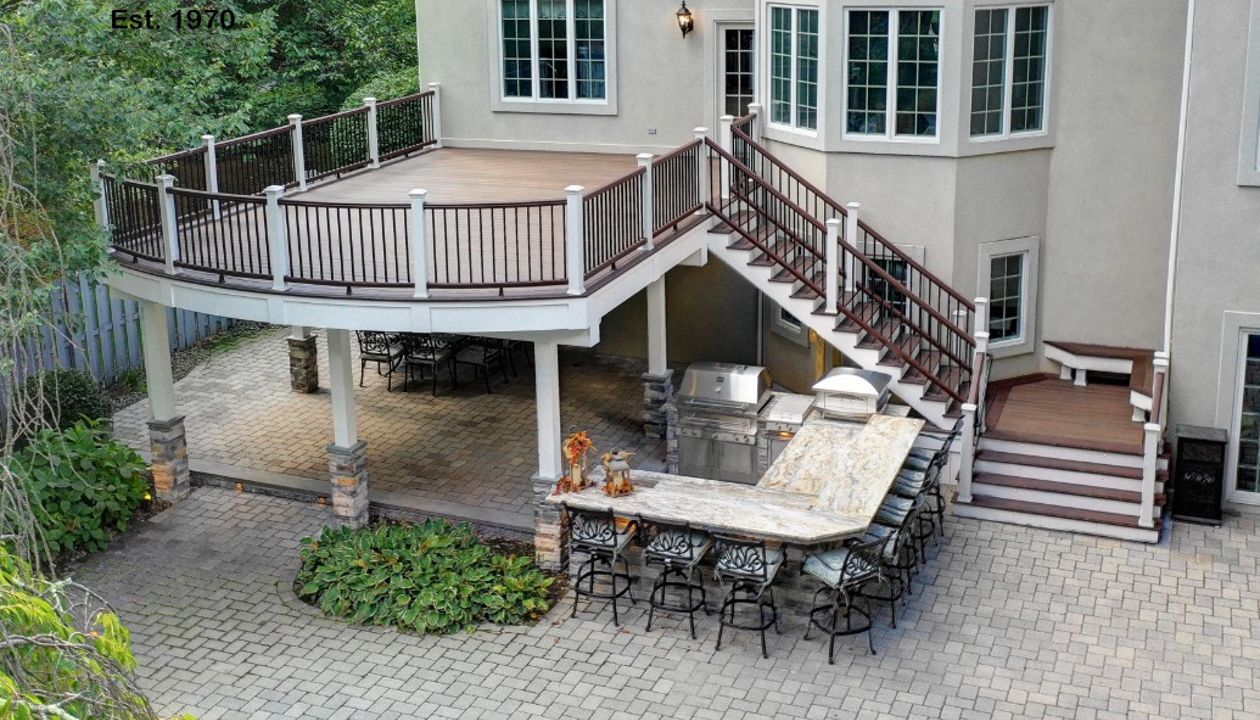 Curved edge deck using Trex Transcend Spiced Rum decking with Vintage Lantern border and railings between white posts. Also includes a drainage system and ceiling below complete with fans, heaters and speakers.
