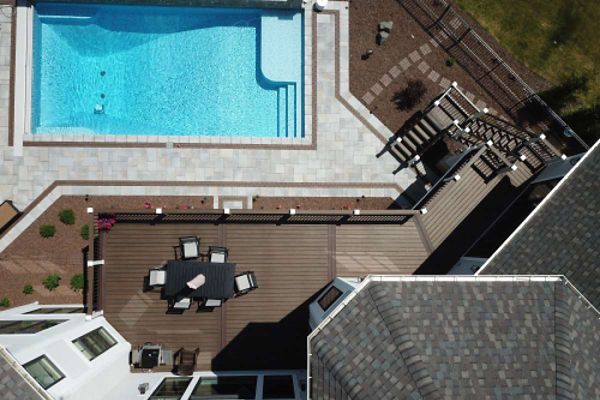 This aerial photograph captures a spacious backyard featuring a rectangular swimming pool with clear blue water and a surrounding stone patio. Adjacent to the pool is a large wooden deck with an outdoor dining table and chairs, partially shaded by an umbrella. The scene includes landscaped garden beds, steps leading to the yard, and the rooftops of a residential home.