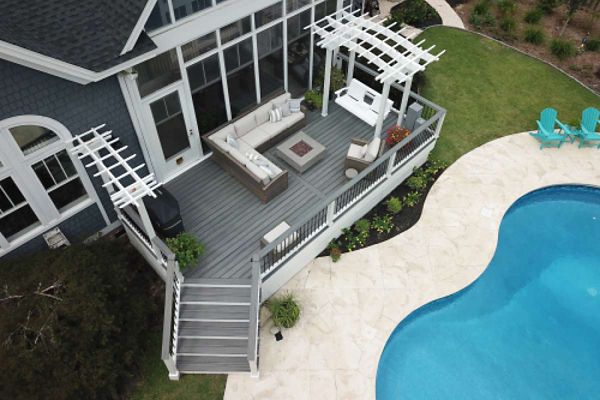Aerial view of a contemporary backyard featuring a gray wooden deck with a sectional sofa and fire pit centerpiece. The deck is attached to a house with large windows and is bordered by white pergolas. Adjacent to the deck is a curved swimming pool with a light stone patio and turquoise lounge chairs, creating a relaxing outdoor setting.