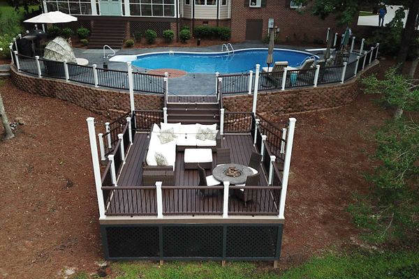 A contemporary outdoor deck features cushioned seating and a fire pit, overlooking a fenced-in swimming pool. The deck is elevated with white and dark railings, set in a landscaped backyard with mulch and mature trees. The pool area includes a brick retaining wall and steps leading up to the house, creating a relaxing and inviting atmosphere.