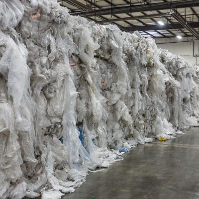 The image shows massive bales of clear and white plastic film stacked along the length of an industrial warehouse. The setting is indoors, with a concrete floor and high ceilings featuring exposed beams and fluorescent lighting. No people are visible, and the mood is utilitarian, emphasizing recycling or waste management. The scene is dominated by the texture and volume of the compressed plastic material.